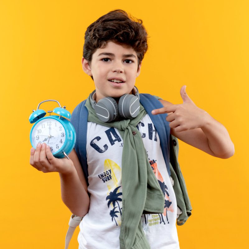 pleased-little-schoolboy-wearing-back-bag-headphones-holding-points-alarm-clock-isolated-yellow-background
